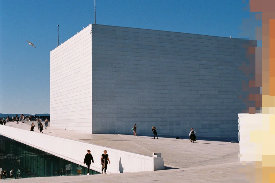 Folk som spaserer på det hvite marmortaket på Operaen i Oslo en solrik dag, med scenetårnet bak og åsene i det fjerne